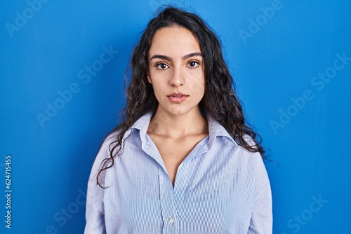 Young brunette woman standing over blue background relaxed with serious expression on face. simple and natural looking at the camera.