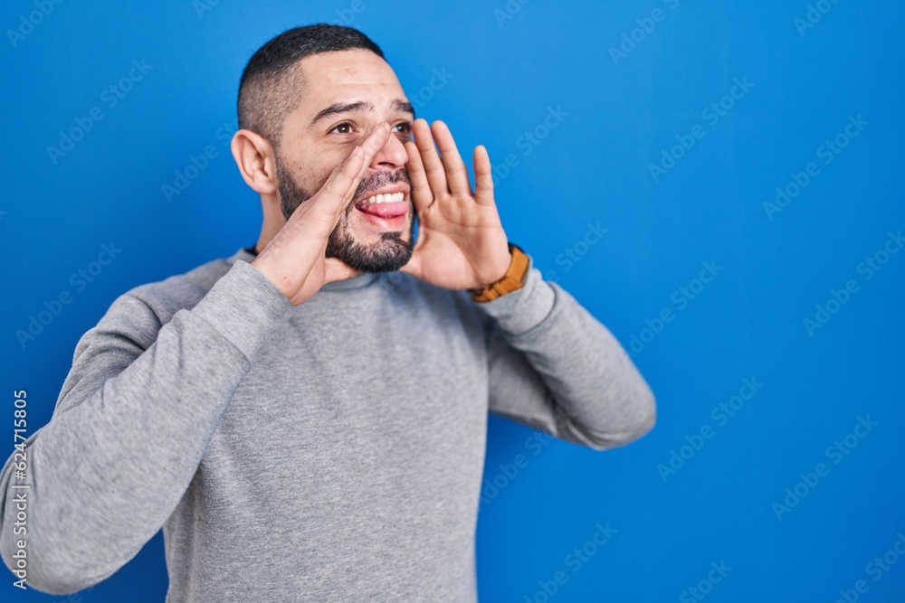 Hispanic man standing over blue background shouting and screaming loud to side with hand on mouth. communication concept. Hispanic man standing over blue background shouting and screaming loud to side with hand on mouth. communication concept.