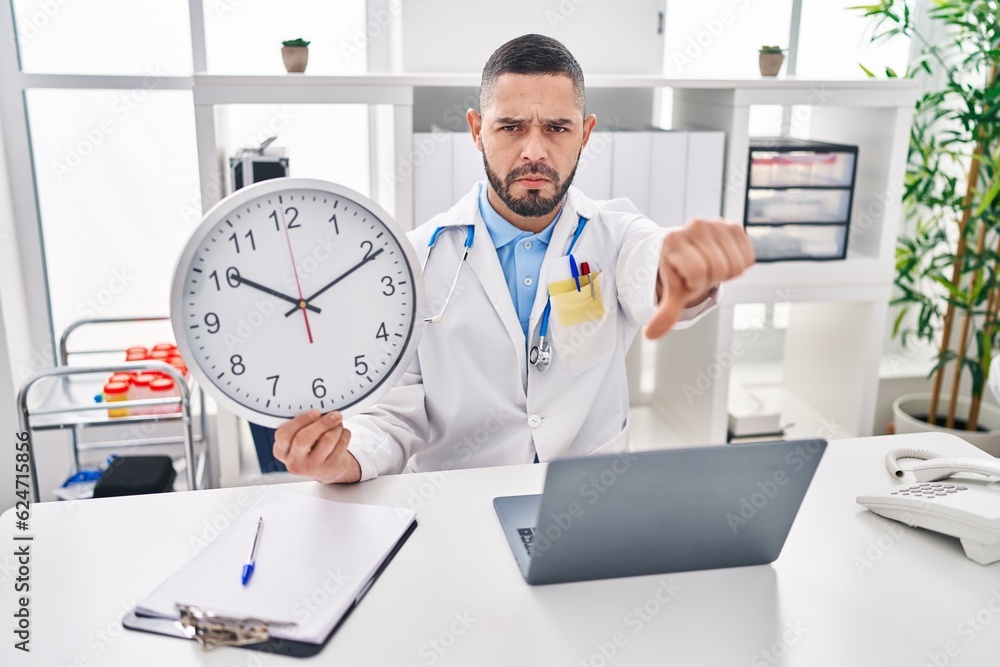 Hispanic doctor man holding clock at the clinic with angry face, negative sign showing dislike with thumbs down, rejection concept Hispanic doctor man holding clock at the clinic with angry face, negative sign showing dislike with thumbs down, rejection concept