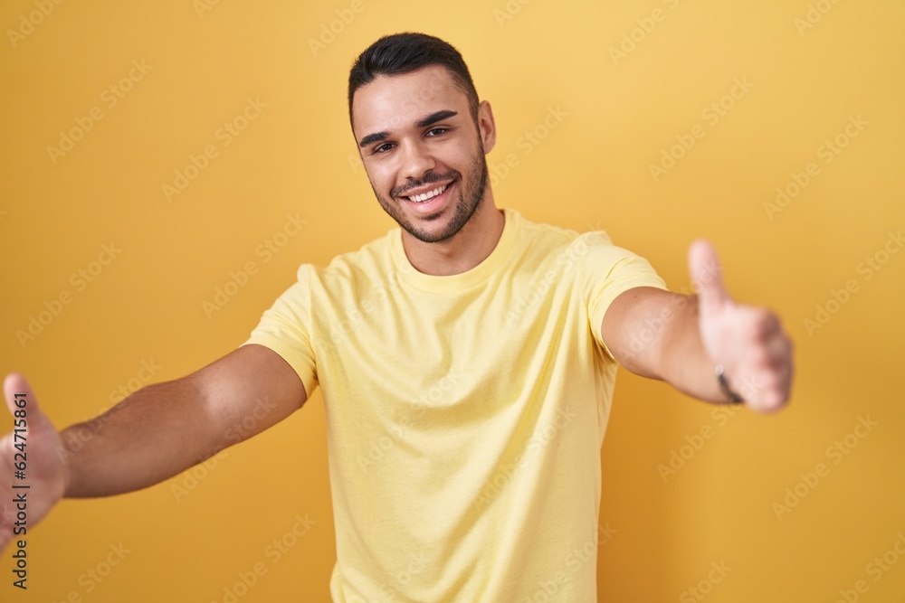 Young hispanic man standing over yellow background looking at the camera smiling with open arms for hug. cheerful expression embracing happiness. Young hispanic man standing over yellow background looking at the camera smiling with open arms for hug. cheerful expression embracing happiness.