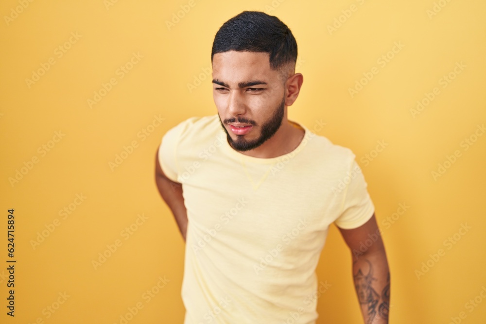 Young hispanic man standing over yellow background suffering of backache, touching back with hand, muscular pain Young hispanic man standing over yellow background suffering of backache, touching back with hand, muscular pain