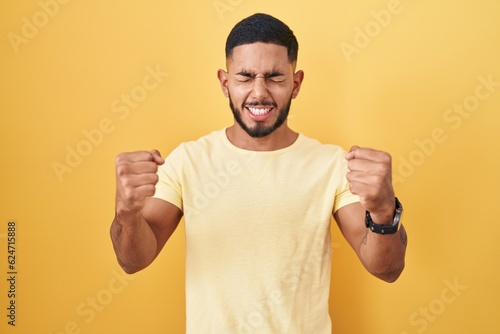 Young hispanic man standing over yellow background excited for success with arms raised and eyes closed celebrating victory smiling. winner concept.