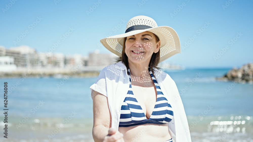 Middle age hispanic woman tourist smiling wearing bikini and summer hat at the beach