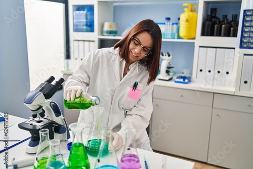 Young beautiful hispanic woman scientist smiling confident pouring liquid on test tube at laboratory