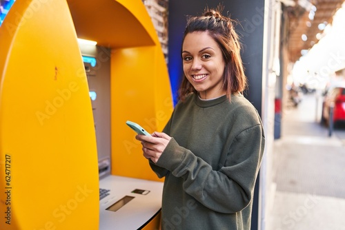 Valokuva Young beautiful hispanic woman using smartphone standing by bank teller at stree