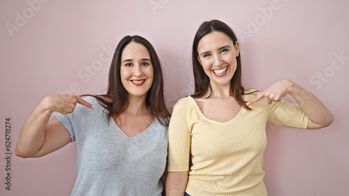 Two women smiling confident pointing with finger to herself over isolated pink background