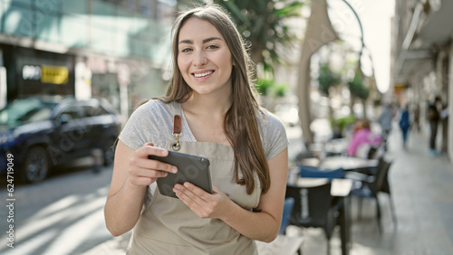 Young beautiful hispanic woman waitress smiling confident using touchpad at coffee shop terrace