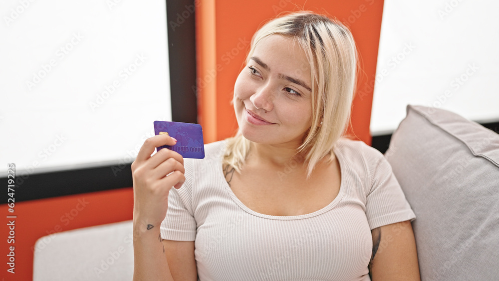 Young beautiful hispanic woman holding credit card sitting on sofa thinking at home