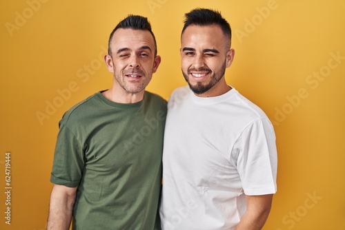 Homosexual couple standing over yellow background winking looking at the camera with sexy expression, cheerful and happy face.