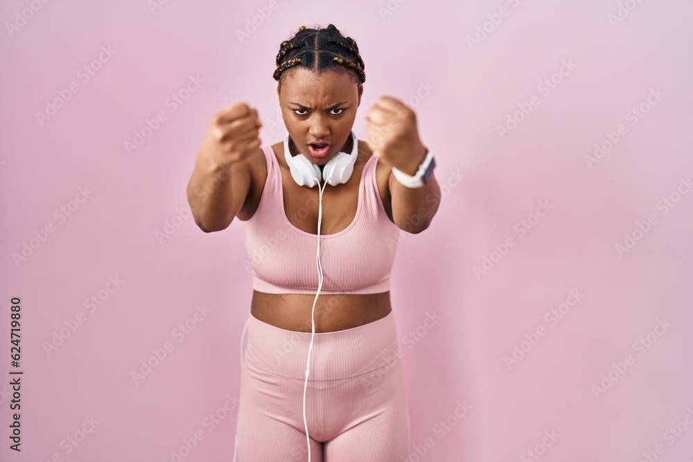 African american woman with braids wearing sportswear and headphones angry and mad raising fists frustrated and furious while shouting with anger. rage and aggressive concept.
