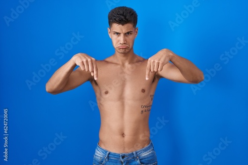 Young hispanic man standing shirtless over blue background pointing down looking sad and upset, indicating direction with fingers, unhappy and depressed.