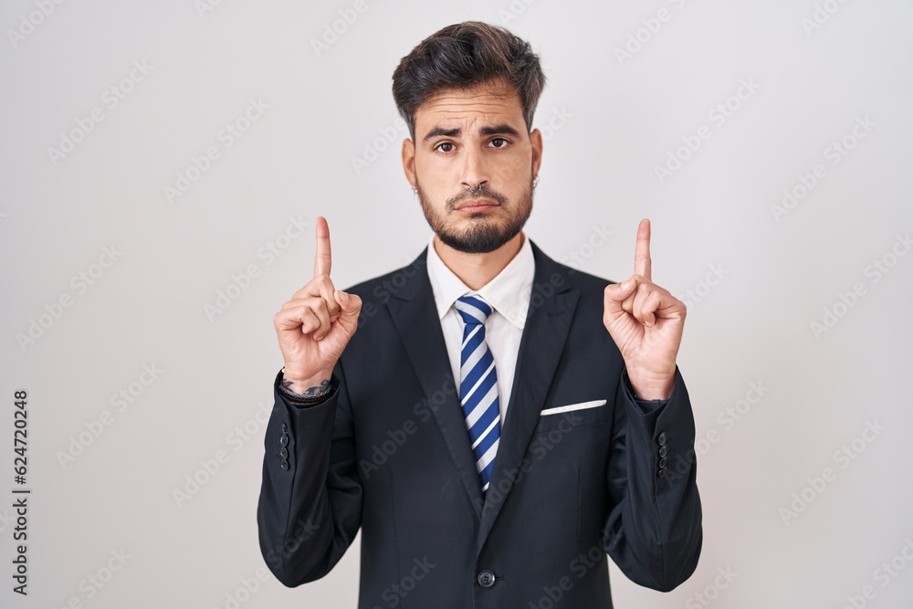 Young hispanic man with tattoos wearing business suit and tie pointing up looking sad and upset, indicating direction with fingers, unhappy and depressed. Young hispanic man with tattoos wearing business suit and tie pointing up looking sad and upset, indicating direction with fingers, unhappy and depressed.