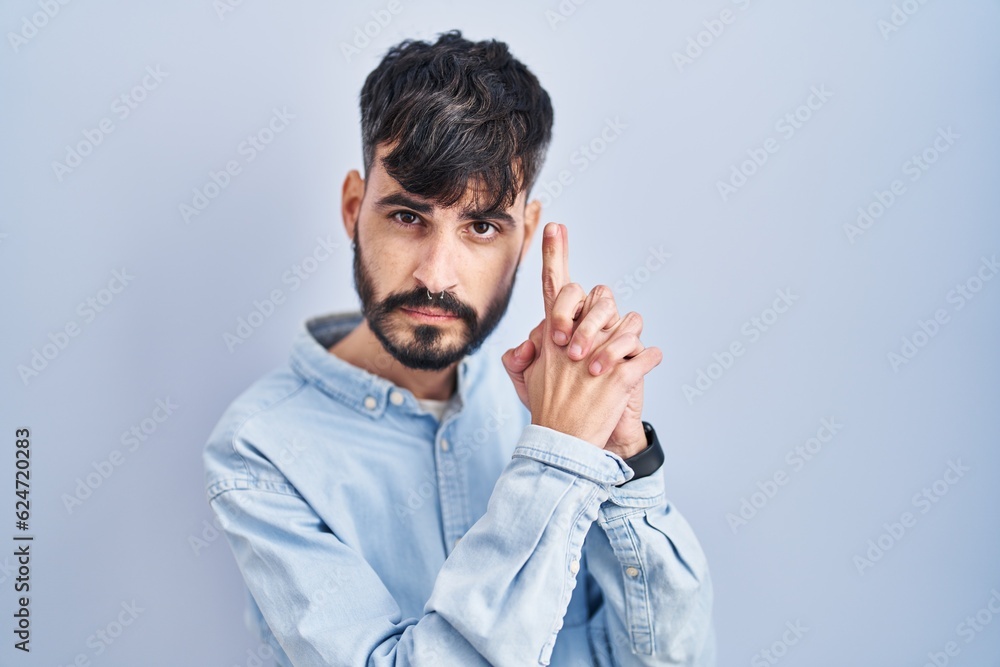 Young hispanic man with beard standing over blue background holding symbolic gun with hand gesture, playing killing shooting weapons, angry face Young hispanic man with beard standing over blue background holding symbolic gun with hand gesture, playing killing shooting weapons, angry face
