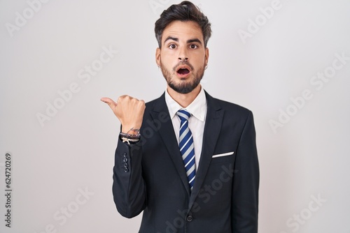 Young hispanic man with tattoos wearing business suit and tie surprised pointing with hand finger to the side, open mouth amazed expression.