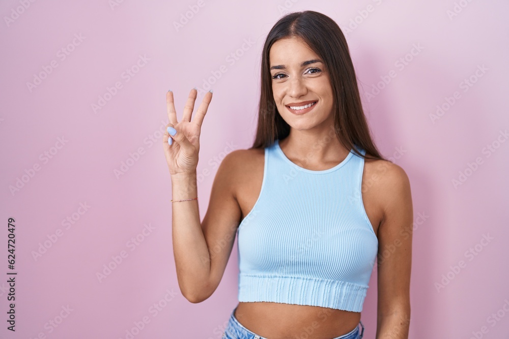 Young brunette woman standing over pink background showing and pointing up with fingers number three while smiling confident and happy.