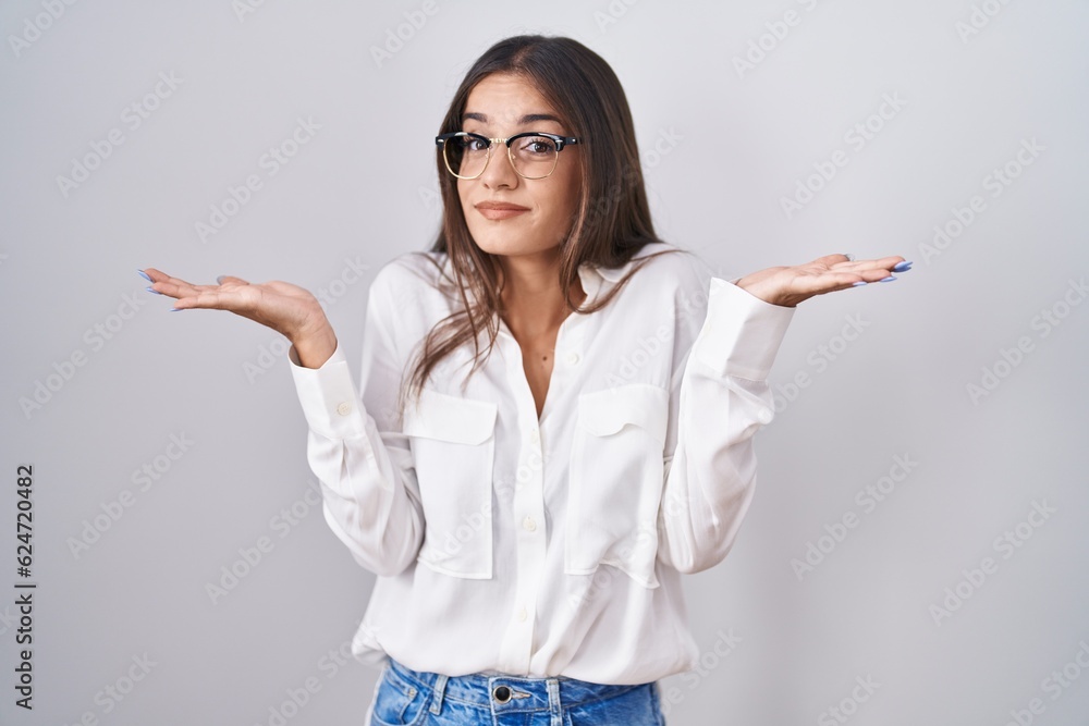 Young brunette woman wearing glasses clueless and confused expression with arms and hands raised. doubt concept. Young brunette woman wearing glasses clueless and confused expression with arms and hands raised. doubt concept.