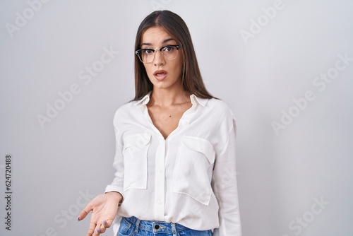 Young brunette woman wearing glasses in shock face, looking skeptical and sarcastic, surprised with open mouth