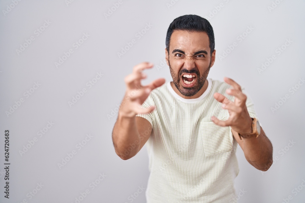 Fototapeta premium Hispanic man with beard standing over isolated background shouting frustrated with rage, hands trying to strangle, yelling mad