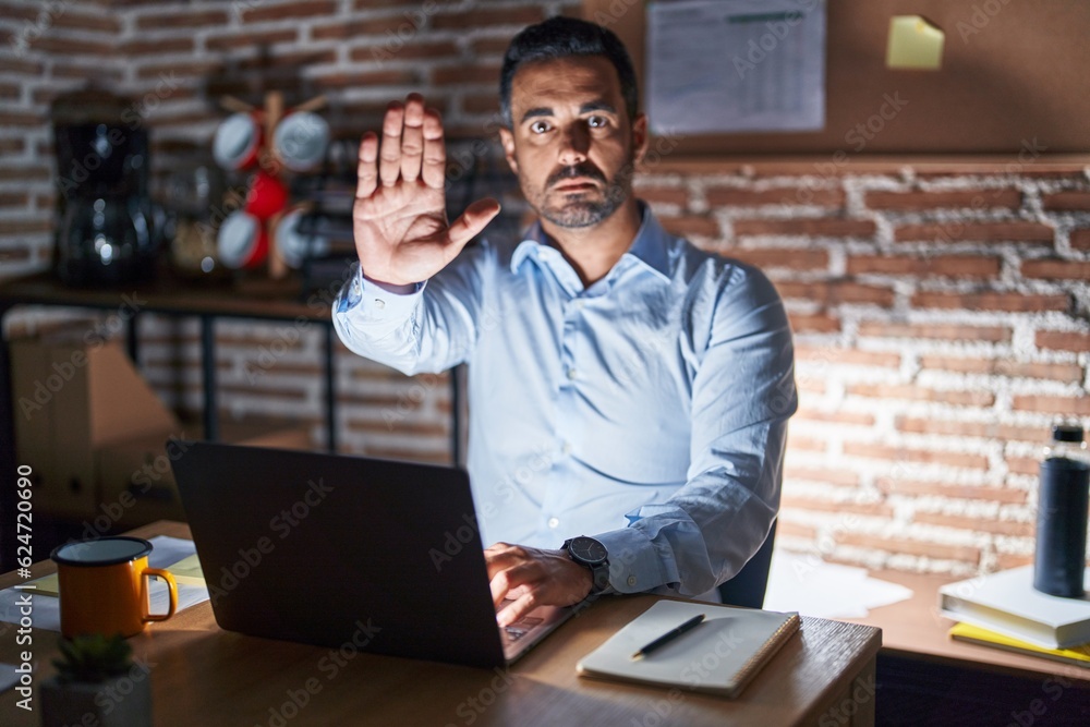 Hispanic man with beard working at the office at night doing stop sing with palm of the hand. warning expression with negative and serious gesture on the face.