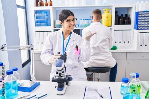 Young woman working at scientist laboratory pointing thumb up to the side smiling happy with open mouth