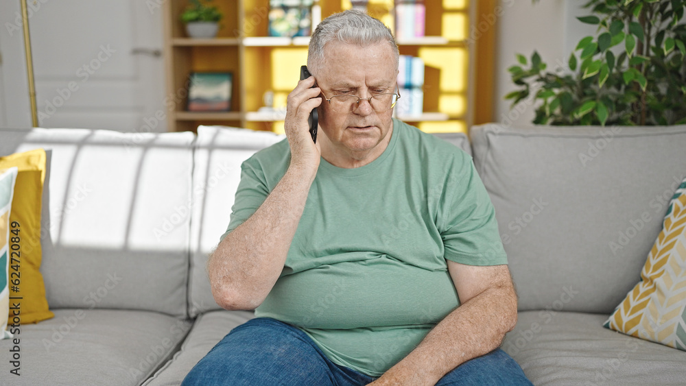Middle age grey-haired man speaking on the phone sitting on sofa at home