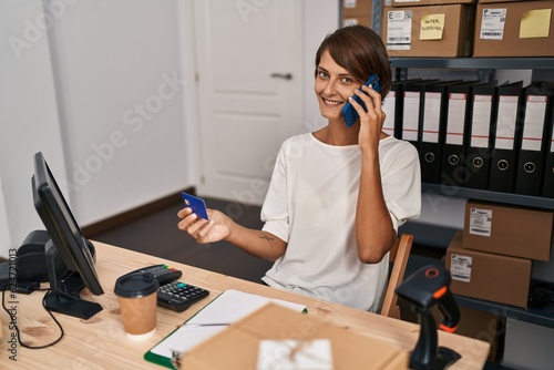 Young beautiful hispanic woman ecommerce business worker talking on smartphone holding credit card at office