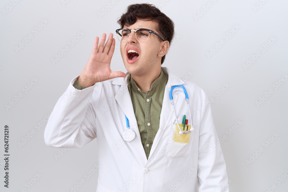 Young non binary man wearing doctor uniform and stethoscope shouting and screaming loud to side with hand on mouth. communication concept. Young non binary man wearing doctor uniform and stethoscope shouting and screaming loud to side with hand on mouth. communication concept.