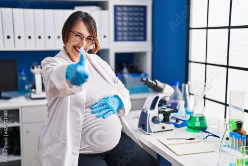 Pregnant woman working at scientist laboratory approving doing positive gesture with hand  thumbs up smiling and happy for success. winner gesture.