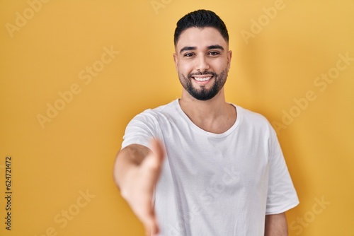 Young handsome man wearing casual t shirt over yellow background smiling friendly offering handshake as greeting and welcoming. successful business.