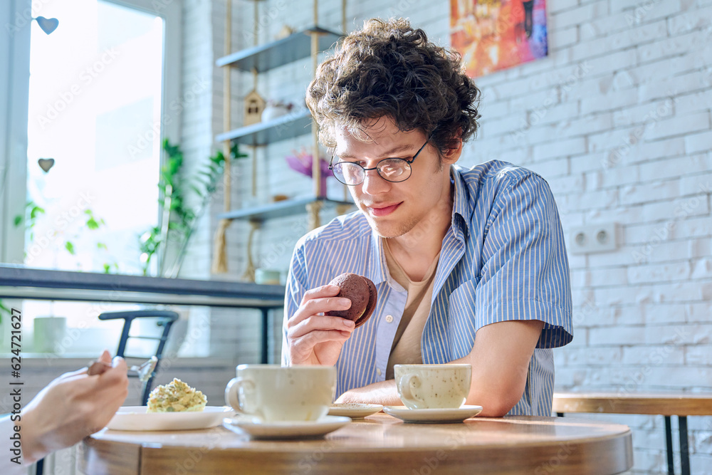 Young guy sitting at table in coffee shop drinks coffee and eats cake Young guy sitting at table in coffee shop drinks coffee and eats cake