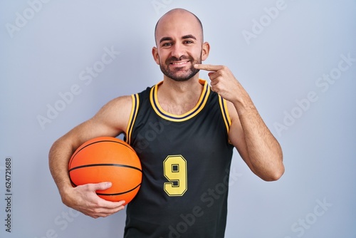 Young bald man with beard wearing basketball uniform holding ball smiling cheerful showing and pointing with fingers teeth and mouth. dental health concept.
