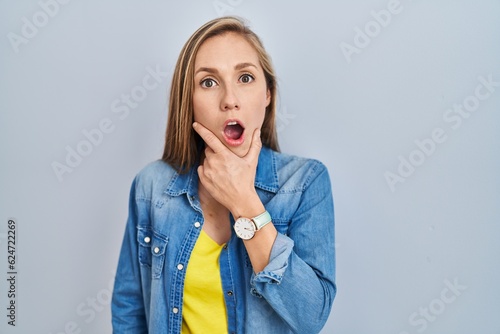 Konstfotografi Young blonde woman standing over blue background looking fascinated with disbeli