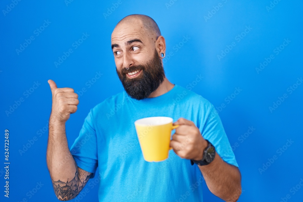 Young hispanic man with beard and tattoos drinking a cup of coffee smiling with happy face looking and pointing to the side with thumb up. Young hispanic man with beard and tattoos drinking a cup of coffee smiling with happy face looking and pointing to the side with thumb up.
