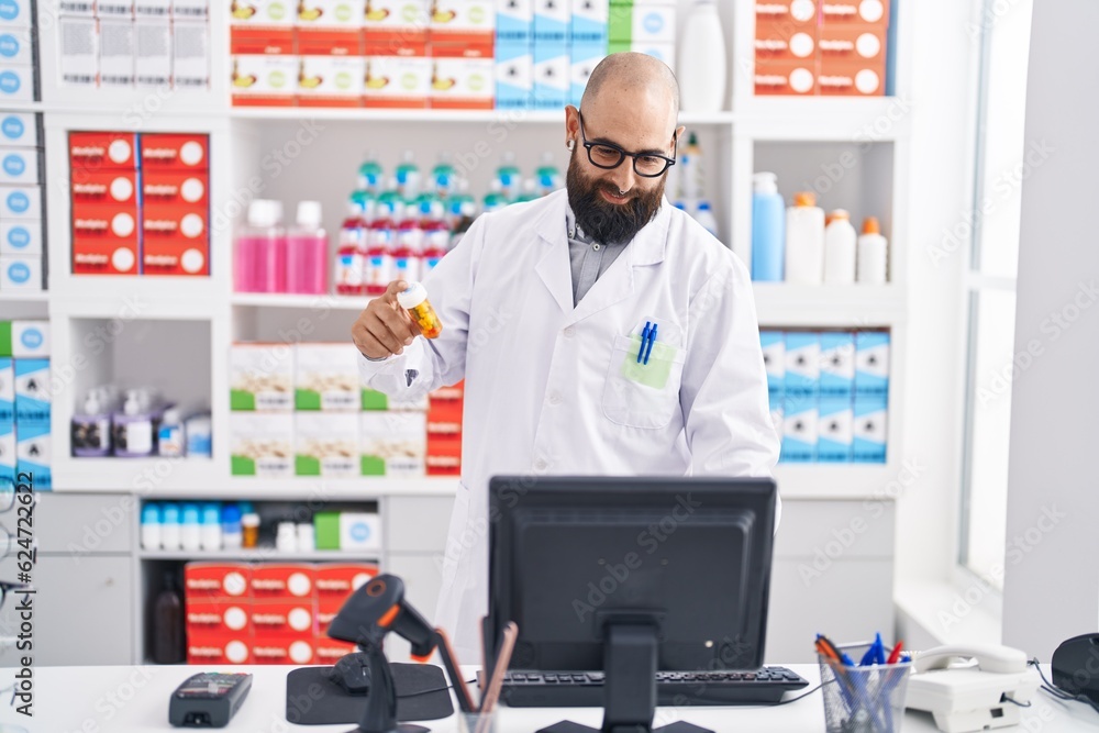 Young bald man pharmacist using computer holding pills bottle at pharmacy