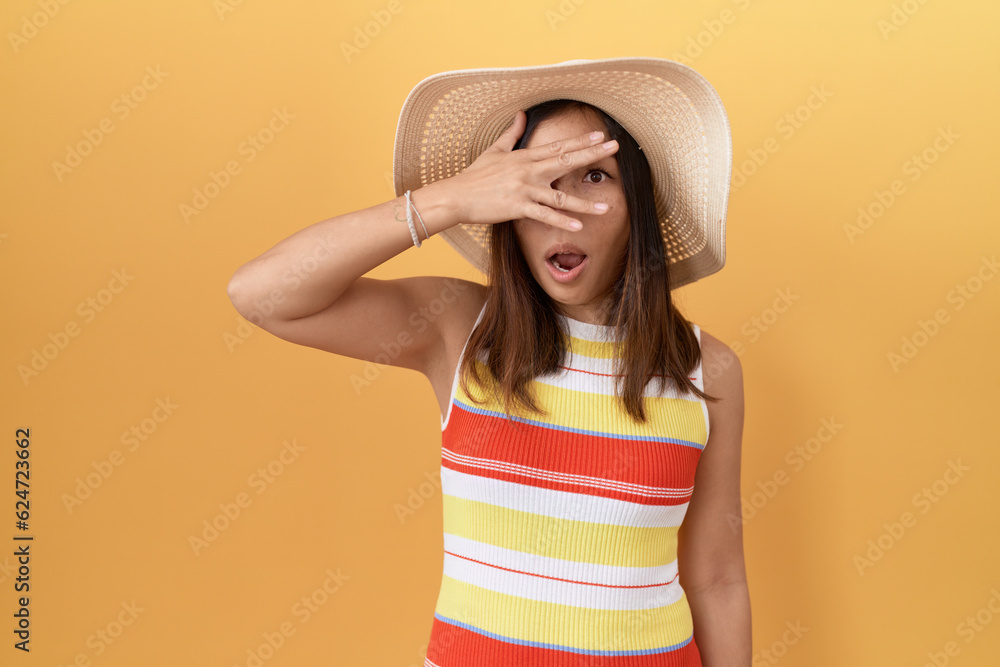 Middle age chinese woman wearing summer hat over yellow background peeking in shock covering face and eyes with hand, looking through fingers with embarrassed expression. Middle age chinese woman wearing summer hat over yellow background peeking in shock covering face and eyes with hand, looking through fingers with embarrassed expression.