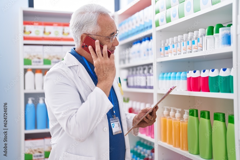 Middle age grey-haired man pharmacist talking on smartphone reading document at pharmacy
