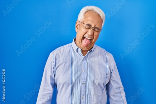 Hispanic senior man wearing glasses sticking tongue out happy with funny expression. emotion concept.