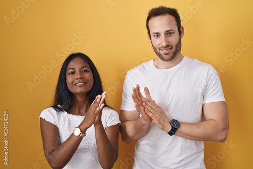 Interracial couple standing over yellow background clapping and applauding happy and joyful, smiling proud hands together