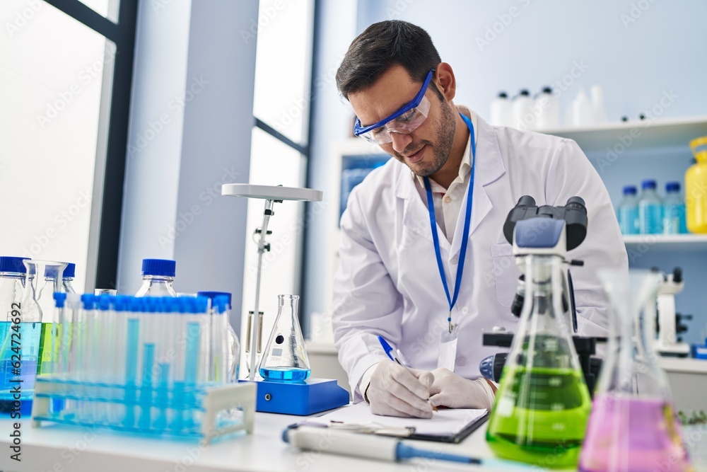 Fototapeta premium Young hispanic man scientist measuring liquid writing report at laboratory
