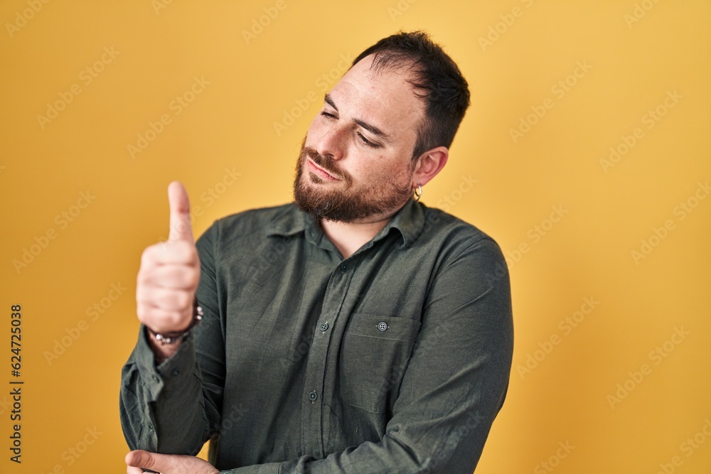 Plus size hispanic man with beard standing over yellow background looking proud, smiling doing thumbs up gesture to the side Plus size hispanic man with beard standing over yellow background looking proud, smiling doing thumbs up gesture to the side