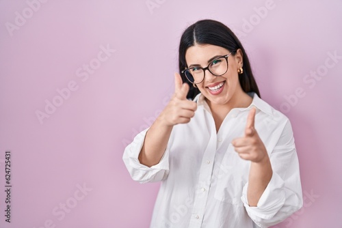 Young brunette woman standing over pink background pointing fingers to camera with happy and funny face. good energy and vibes.