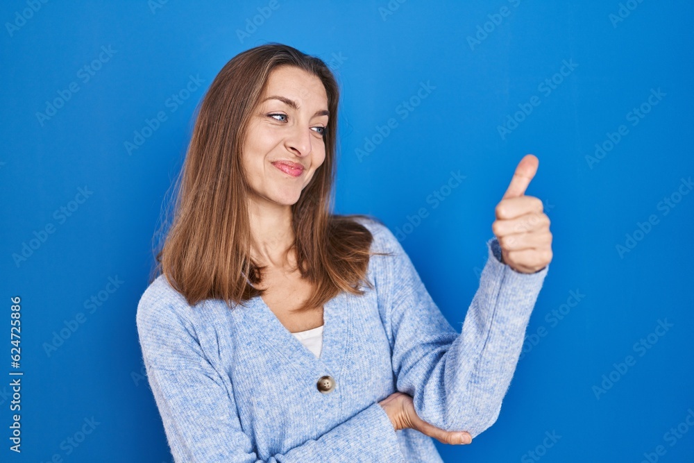 Young woman standing over blue background looking proud, smiling doing thumbs up gesture to the side Young woman standing over blue background looking proud, smiling doing thumbs up gesture to the side