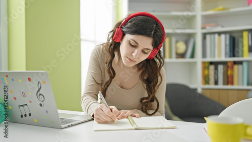 Young beautiful hispanic woman student using laptop and headphones writing on notebook at library university