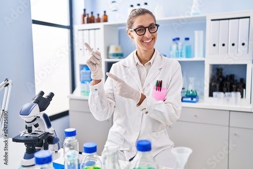 Young woman working at scientist laboratory smiling and looking at the camera pointing with two hands and fingers to the side.