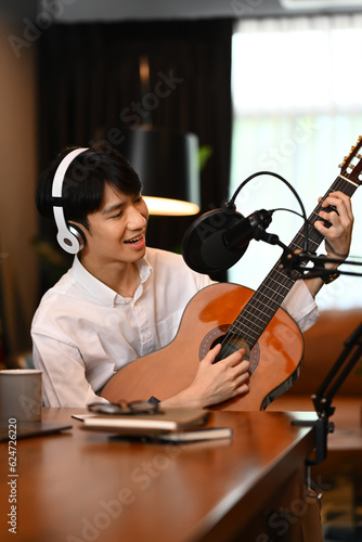 Happy young asian in wireless headphones playing acoustic guitar while recording podcast near microphone in home studio