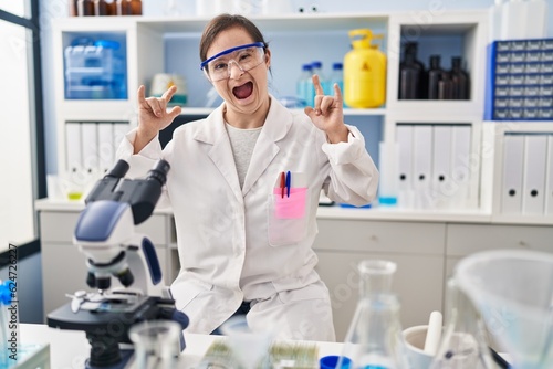 Hispanic girl with down syndrome working at scientist laboratory shouting with crazy expression doing rock symbol with hands up. music star. heavy music concept.