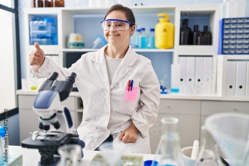Hispanic girl with down syndrome working at scientist laboratory looking proud, smiling doing thumbs up gesture to the side