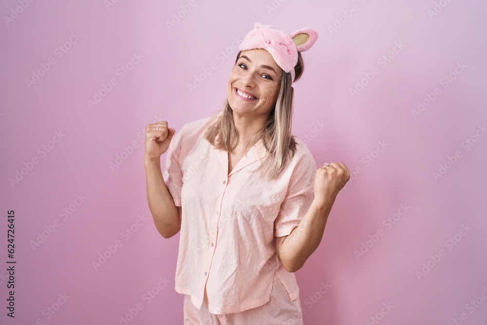 Blonde caucasian woman wearing sleep mask and pajama very happy and excited doing winner gesture with arms raised, smiling and screaming for success. celebration concept. Blonde caucasian woman wearing sleep mask and pajama very happy and excited doing winner gesture with arms raised, smiling and screaming for success. celebration concept.