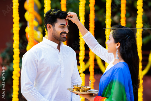 Indian girl with puja plate applying tilak to husband before playing holi festival o flower decorated background - concept of festive rituals, traditional and culture and celebration photo