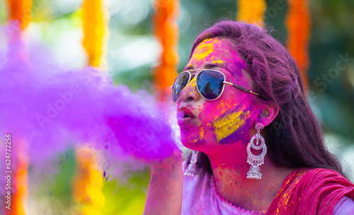Young indian girl with sunglasses blowing holi colour powder from hand during on flower decoration background - concept of holi festival celebration, Indian tradition and colourful culture